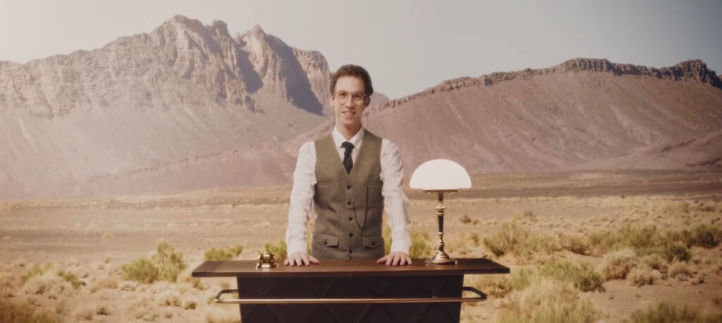 Concierge in Weste steht hinter einem Pult mit Lampe in einer Wüstenlandschaft, Standbild aus dem Werbefilm Bosch Roadside Assistance