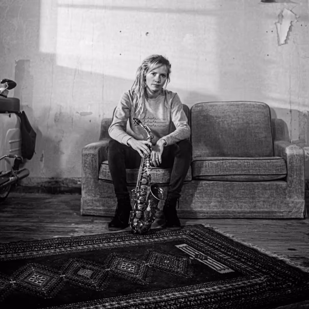 Eine Person mit Dreadlocks sitzt auf einem grauen Sofa und hält ein Saxophon. Der Boden ist mit einem gemusterten Teppich bedeckt, in einem Raum mit abblätternder Wandfarbe. Das Bild ist in schwarz-weiß.