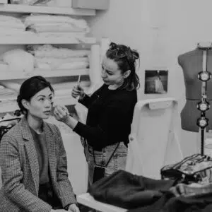 Videoproduktion Make-up-Künstlerin trägt schwarze Kleidung und Jeans, schminkt sitzende Person mit kariertem Blazer in einem Raum mit Regalen voller Stoffe und Schneiderpuppe.