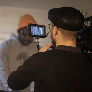 Videoproduktion Kameramann mit schwarzer Lederkappe und Brille bedient eine Filmkamera bei Dreharbeiten, im Hintergrund unscharfe Person mit beiger Mütze und hellgrauem Hoodie.