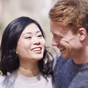 Eine männlich und eine weiblich gelesene Person schauen sich an. Beide tragen lässige Kleidung und lächeln, während sie sich an einem sonnigen Ort befinden.