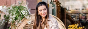 Eine Person mit langen, braunen Haaren sitzt auf einem geflochtenen Stuhl in einem Café. Im Hintergrund sind Blumen und Pflanzen sichtbar.