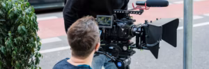 Ein Kameramann filmt mit einer professionellen Kamera auf einem Stativ. Im Vordergrund ist der Rücken des Kameramanns zu sehen, der auf die Kamera blickt. Die Kamera hat ein großes Objektiv und ein externes Monitor-Display.