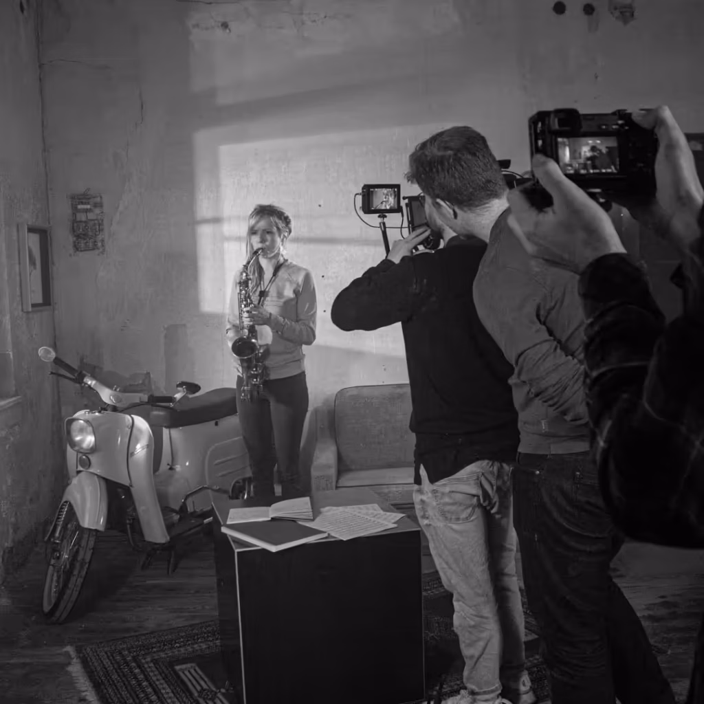 Zwei Kameraleute filmen eine Person mit Saxophon in einem Raum mit Sofa und weißem Moped, Schwarzweißfoto
