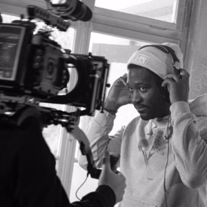 Eine Person trägt einen grauen Kapuzenpullover von der Marke sofalich und Kopfhörer, während sie mit einer Filmkamera gefilmt wird. Die Kamera wird von der filmenden Person auf den Schultern getragen.